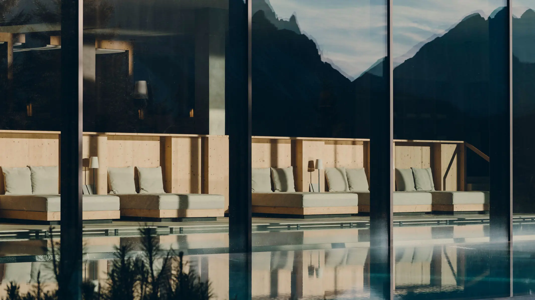 The indoor pool seen through its glass wall, a row of timber-framed loungers along the water with the mountain ridge reflected across both the glazing and the surface.