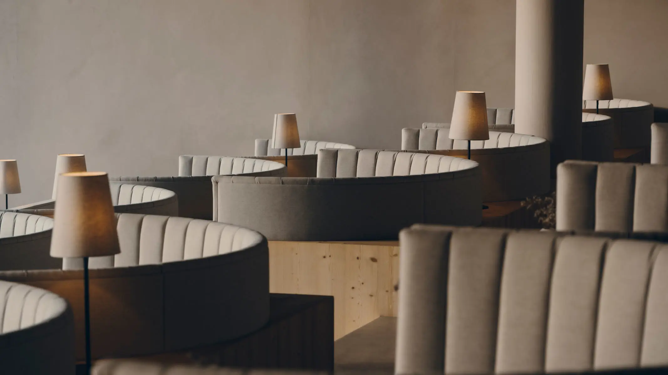 Curved fluted banquettes in pale fabric stepping back through the dining room, each with a slim cordless table lamp and a small vase of dried stems.