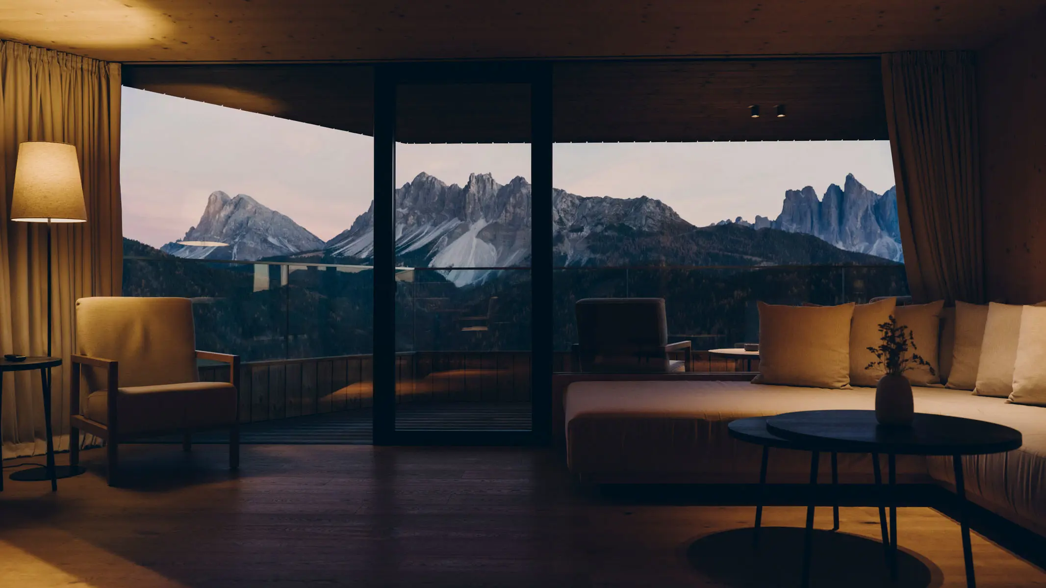 A suite sitting room at dusk, sliding glass onto a balcony with the pale Dolomite skyline beyond, a long cushioned daybed, a leather armchair and a round black table on dark timber floor.