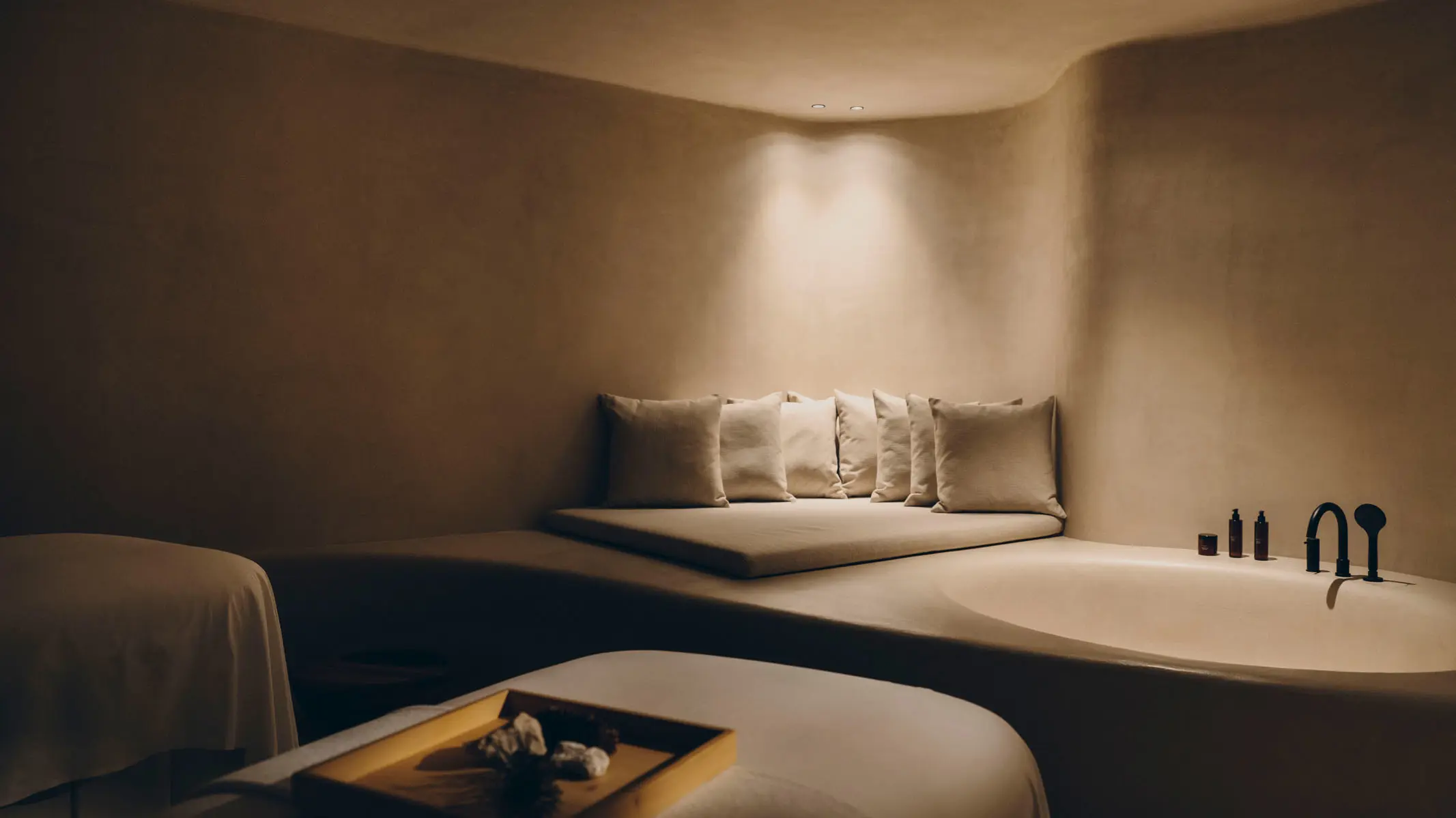 A treatment room moulded in seamless clay-coloured plaster, a curved bench of pillows in the corner, a sunken oval bath with black tapware and two draped massage tables.