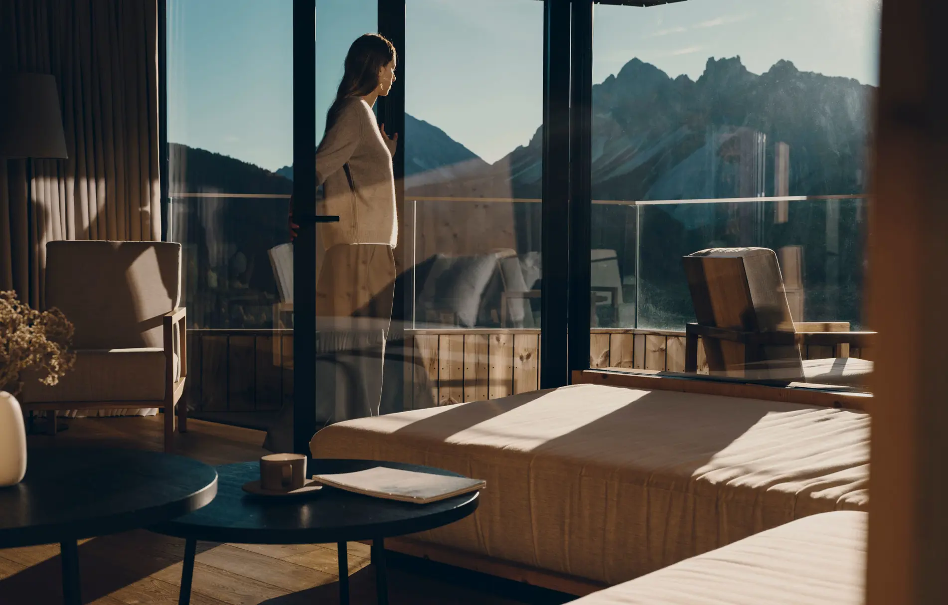 Morning in a room whose glass corner opens onto jagged Dolomite peaks, a guest standing at the sliding door in a robe, coffee and a book on the low table behind her.