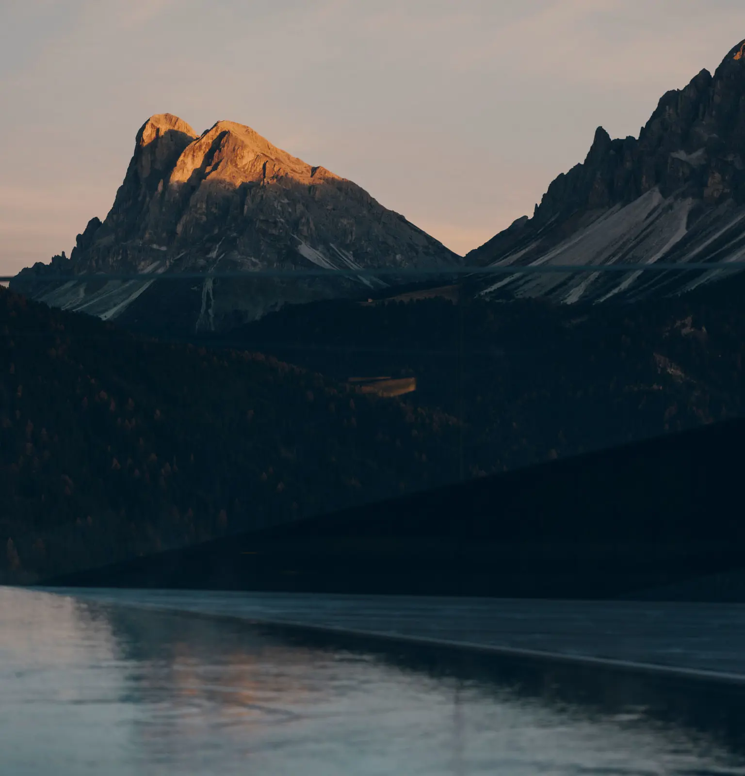 The lip of the outdoor pool at first light, still water running to a rimless edge with the Sass de Putia massif beyond, its summit catching orange sun above ridges still in shadow.