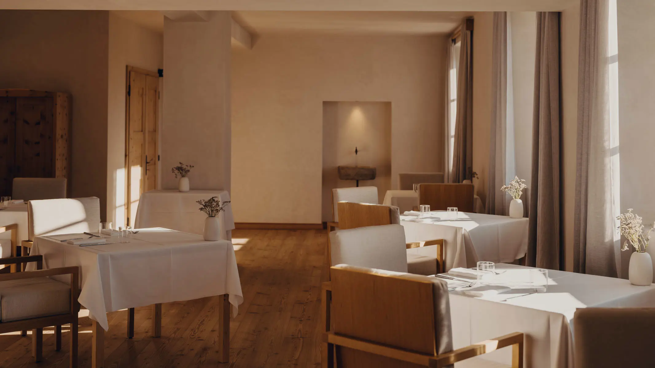 The restaurant in daylight, white-clothed tables with pale timber chairs spread across a wide-plank floor, sun falling through tall curtained windows onto lime-plastered walls.