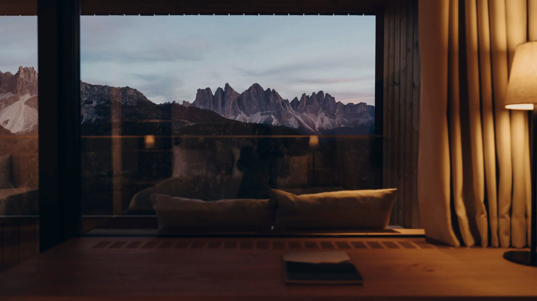 A cushioned window seat looking out at the peaks through a single large pane, a linen curtain drawn back at one side and an open notebook on the timber ledge in front.