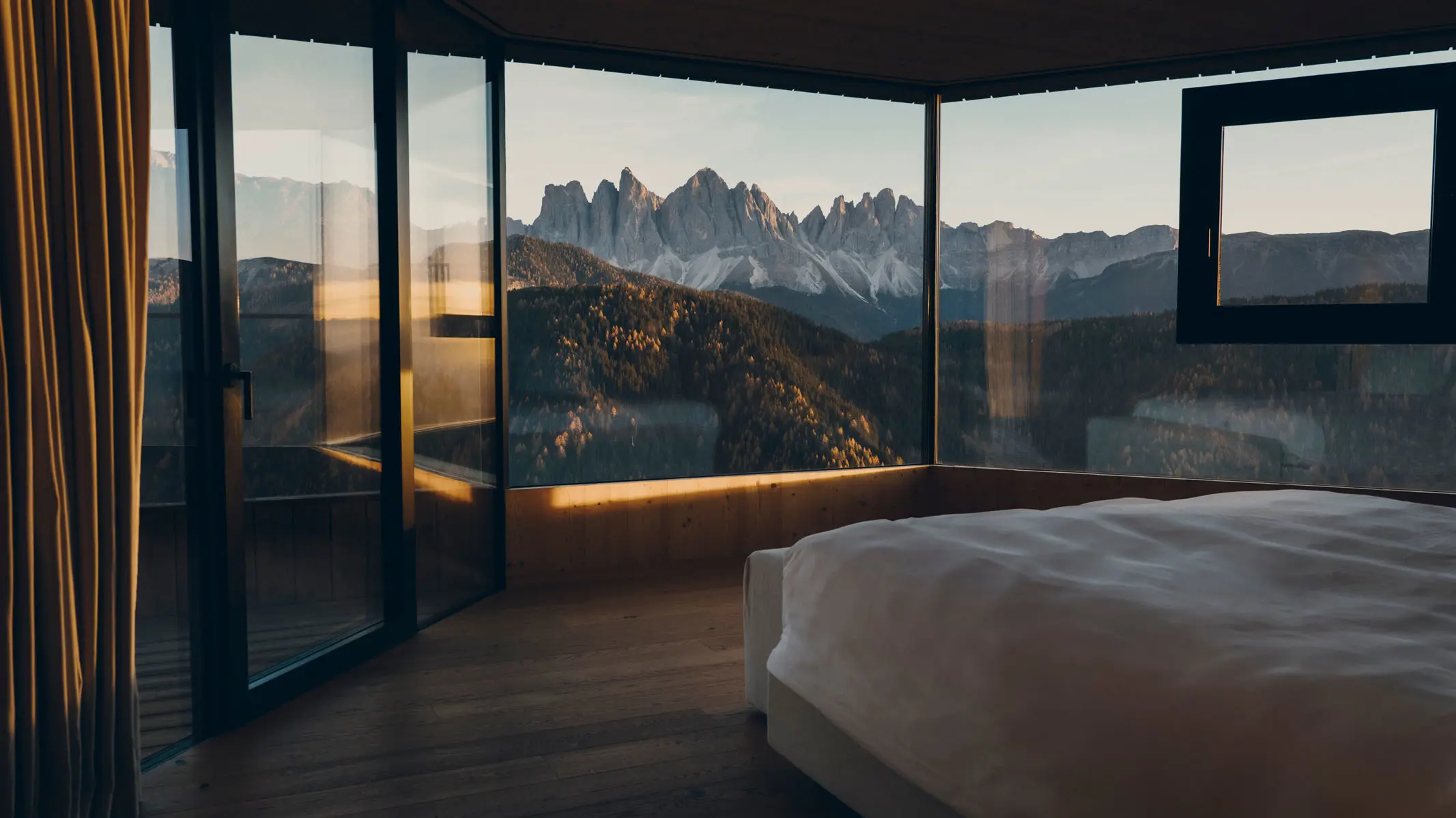 A bed set against a corner of full-height glass, the Odle peaks and a forested ridge filling both walls, with a timber-lined ceiling and a strip of warm light along the sill.
